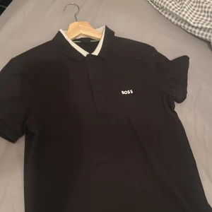 Svart pikétröja från BOSS - Snygg svart pikétröja från BOSS med vit logga på bröstet och ribbad krage i svart och vitt. Tröjan har tre knappar framtill och korta ärmar. Perfekt för dig som gillar stilrena och klassiska plagg med premiumkänsla. Två små håll finns vid bossen märker 