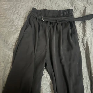 Svarta vida byxor med knytband - Säljer ett par svarta byxor med hög midja och vida ben. Byxorna har ett snyggt knytband i midjan med metallring och papperspåse-liknande midjedetalj. Perfekta för en chill och stilren look. Storlek L. Fint skick! 