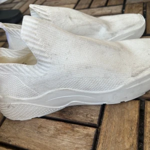 Vita slip-on sneakers i stickad mesh - Säljer ett par helvita slip-on sneakers med chunky sula och stickad ovandel i mesh. Skorna har en sportig vibe och är superlätta att dra på tack vare den flexibla, strumpliknande designen. Perfekta för dig som gillar minimalistisk stil och komfort.