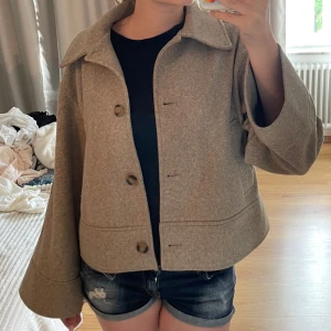 Kappa - Säljer en beige jacka i ull med bred krage och stora knappar framtill. Jackan har en boxig, loose passform och vida ärmar som ger en chill vibe. Perfekt för lager-på-lager och enkel att styla till jeans eller kjol.