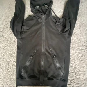 Svart hoodiejacka med goggles C.P. Company - Svart hoodiejacka från C.P. Company med ikoniska goggles i huvan. Jackan har dragkedja framtill, två fickor med dragkedja och justerbar huva. Perfekt för dig som vill sticka ut med streetwear-vibe och coola detaljer. Byta funkar!