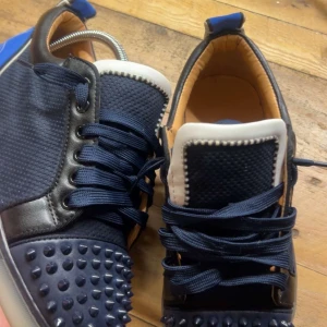 Mörkblåa Louboutins med spikes - Unika sneakers i mörkblått med coola nitar på tån, vita detaljer och blå häl med stor Louboutin-liknande logga. Skorna har snörning, rund tå och platt sula. Materialmix av syntet och skinnimitation, perfekt för dig som vill sticka ut. Priset går att diskutera
