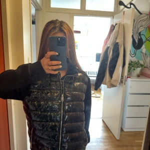 Svart dunväst från Moncler - Säljer en svart dunväst från Moncler med glansig finish och quiltad design. Västen har dragkedja framtill och klassisk Moncler-logga på bröstet. Insidan har ett coolt serietidningsmönster och västen är perfekt för lager-på-lager-stil. Har dock inte bevis på att den är äkta då jag fick av min vän.