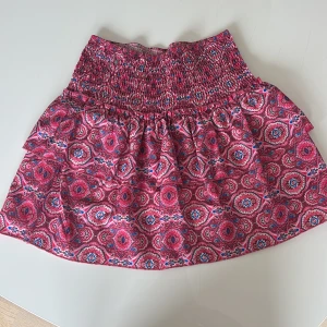 Rosa mönstrad kortkjol med volang - Superfin kortkjol i rosa med färgglatt mönster, bred smock i midjan. Kjolen har volang och inbyggda shorts under för extra komfort. Perfekt för dig som gillar färg och vill sticka ut lite extra i sommar. Helt ny och aldrig använd