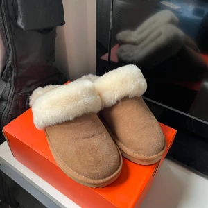 Beiga fluffiga tofflor från Cedrico - Supermysiga tofflor från Cedrico i beige mocka med fluffig vit päls runt hela öppningen och insidan. Perfekta för att hålla fötterna varma hemma, med en platt sula i gummi som ger bra grepp. Klassisk slip-in modell med rund tå.