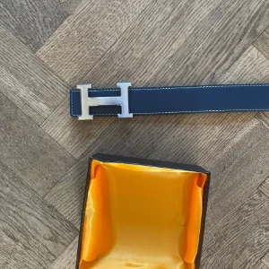 H bälte - Säljer ett svart skärp från H med ikoniskt H-spänne i silverfärgad metall. Bältet är i slätt skinn med vita kontrastsömmar och kommer i originalask med orange insida. Perfekt accessoar för att lyfta din outfit.