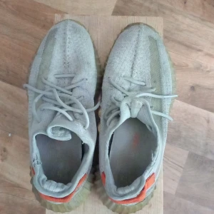 Adidas Yeezy Boost 350 V2 ljusgrå - Adidas Yeezy Boost 350 V2 i ljusgrått med stickad ovandel och genomskinlig sula. Skorna har snörning och en orange detalj på hälen. Ikonisk siluett med platt sula och rund tå, perfekt för dig som gillar streetwear.