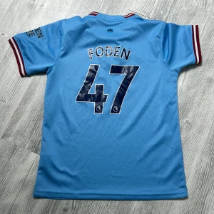 Manchester City Foden 47 matchtröja - Ljusblå Manchester City fotbollströja med Foden och nummer 47 på ryggen. Kortärmad modell med klubbmärke och Etihad Airways-logga framtill. Röd och vit rand på ärmslut och krage. Tillverkad i lätt och ventilerande material, perfekt för fotboll eller supporterliv.