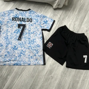 Portugal Ronaldo #7 fotbollströja set - Snyggt Portugal fotbollströja och shorts set med Ronaldo och nummer 7. Tröjan är vit med blått grafiskt mönster, svarta detaljer och Portugal-emblem. Matchande svarta shorts med emblem och nummer. Tillverkad i lätt och andningsbart material.