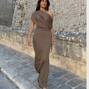Brun långklänning med oneshoulder - Snygg brun långklänning med oneshoulder och draperad överdel. Klänningen har tight passform, kort ärm och slits vid sidan. Tillverkad i mjukt stretchigt material som sitter skönt på kroppen och framhäver siluetten. Perfekt för fest och bröllop.