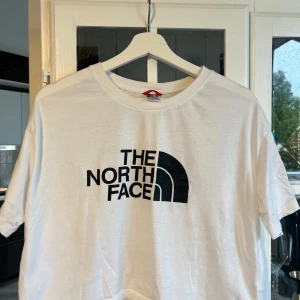 Vit The North Face t-shirt croptop  - Vit t-shirt från The North Face med klassisk svart logga framtill. Modellen är kortärmad och har en rund halsringning. Tillverkad i mjuk bomull, perfekt för en clean och avslappnad stil. Croptop. Storlek medium. 