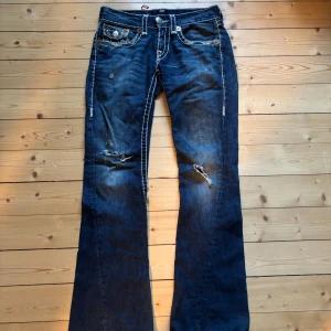 True Religion bootcut jeans blå - Säljer ett par blå jeans från True Religion med tydliga kontrastsömmar, slitningar och bootcut-modell. Jeansen har stora fickor med lock och silverfärgade knappar, samt klassisk True Religion patch bak. Materialet är jeans med en cool, sliten look.