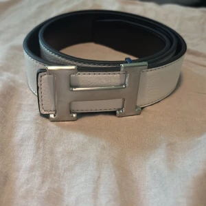 H vändbart skärp med H-spänne - Vit Hrms bälte som även går att vända om man vill till svart. Snyggt vändbart bälte från H med ikoniskt H-spänne i silver. Ena sidan är vit och den andra svart, vilket gör det superenkelt att matcha med olika outfits. Tillverkat i exklusivt skinn med stilren design och bred rem. Perfekt accessoar för att lyfta din look.