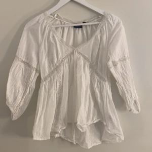 Vit blus med spetsdetalier - Superfin vit blus från Lindex med spetsdetaljer och knyt framtill. Blusen har v-ringning, trekvartsärmar och en luftig, flowy passform. Finns en liten sminkfläck på sidan av armen men inget stort💕