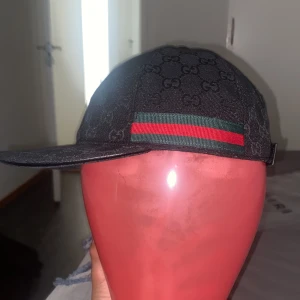 Svart keps med GG-mönster - Svart keps med klassiskt GG-monogram över hela ytan. Kepsen har en böjd skärm, justerbart spänne bak och den ikoniska gröna och röda randen på sidan. Tillverkad i bomullsmix med detaljer i skinn. Perfekt för dig som vill sticka ut med lyxig streetstyle.