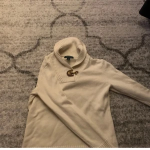 Beige polotröja från Ralph Lauren - Snygg beige polotröja från Lauren Ralph Lauren med ribbad krage och coolt spänne i läder och metall vid halsen. Tröjan är långärmad och gjord i mjuk bomull, perfekt för lager-på-lager under kyliga dagar.