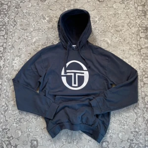 Mörkblå hoodie från Sergio Tacchini XXL  - Snygg mörkblå hoodie från Sergio Tacchini med stort vitt logotryck på bröstet. Tröjan har huva med snörning och är tillverkad i 100% bomull. Perfekt för en chill och sportig stil. XXL men liten i storleken. 