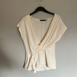Krämvit omlottopp från Zara - Säljer en krämvit omlottopp från Zara med v-ringning och knyt i sidan. Toppen har korta ärmar och är tillverkad i ett mjukt, lätt material som ger ett snyggt fall. Perfekt för dig som gillar stilrena och feminina plagg. Den har tyvärr lite sminkfläckar.