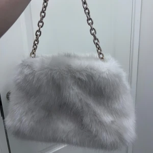 Vit fluffig axelväska med kedja - Supermjuk och fluffig axelväska i vit fuskpäls med en snygg kedjerem i metall. Perfekt för att ge din outfit en trendig och mysig touch. Väskan har en rektangulär form och känns riktigt lyxig att bära.