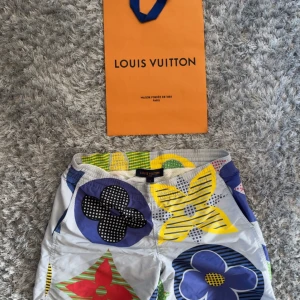 Färgglada Louis Vuitton badshorts S - Unika badshorts från Louis Vuitton med stora blommiga mönster i blått, gult, rött, grönt och svart på ljus bakgrund. Shortsen har elastisk midja och meshfoder för extra komfort. Perfekta för sommaren när du vill sticka ut på stranden.