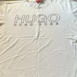 Vit t-shirt från Hugo Boss - Vit t-shirt från Hugo Boss med stor HUGO-logga tryckt framtill i grått. Klassisk rund halsringning och korta ärmar. Tillverkad i mjuk bomull som känns skön mot huden. Perfekt för en clean och stilren look. Storlek Large.