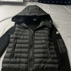 Svart Moncler pufferjacka med huva - Snygg svart pufferjacka från Moncler med stickade ärmar och quiltad front. Jackan har huva, dragkedja framtill och Moncler-logga på ärmen. Perfekt för dig som vill ha en stilren och sportig look med exklusiv känsla.