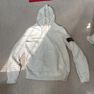 Vit hoodie från Stone Island - Säljer en clean vit hoodie från Stone Island med klassisk patch på vänster ärm. Tröjan har huva med snörning och stor känguruficka framtill. Perfekt för dig som gillar streetwear och vill ha något stilrent i garderoben.