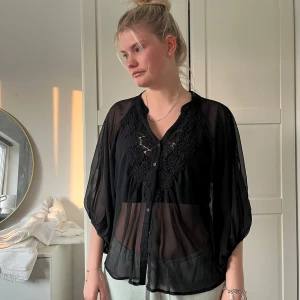 Svart transparent blus med broderi - Säljer en svart, luftig och transparent blus med v-ringning och broderade blommor framtill. Blusen har knappar hela vägen och vida, trekvartsärmar. Perfekt för dig som gillar en chill och trendig look.