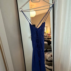 Blå glittrig långklänning med snörning - Snygg blå långklänning med glittriga paljetter och tunna axelband. Klänningen har snörning i ryggen och en draperad urringning framtill. Perfekt för dig som vill sticka ut med en elegant och trendig look.