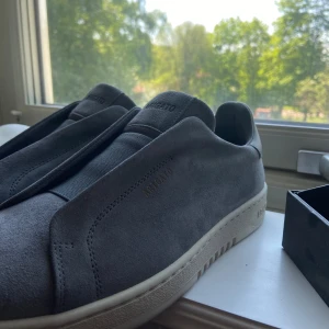 Grå sneakers från Axel Arigato mocka - Nypris 2600kr endast testade! Kartong finnes! Snygga grå sneakers från Axel Arigato i mjuk mocka med minimalistisk design och diskret logga på sidan. Skorna har elastiskt band över vristen istället för klassisk snörning och en vit se sula. Perfekta för dig som gillar stilren och modern look.