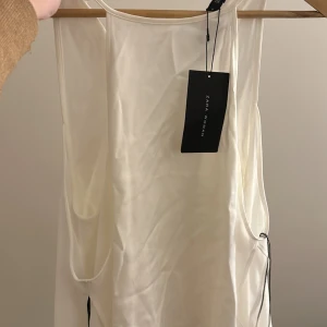 Vit topp  från Zara - Supersnygg vit blus från Zara med v-ringning och ärmlös design. Toppen har en lätt, flowy känsla och är tillverkad i ett tunt, mjukt material som ger en elegant look. Perfekt för varma dagar och stilrena outfits. Säljer denna då den e alldeles för stor på mig. Toppen är långärmad men axelbar jättesnygg aldrig använd, köpt i spanien köpt för ca 428 kr säljer för 200kr.