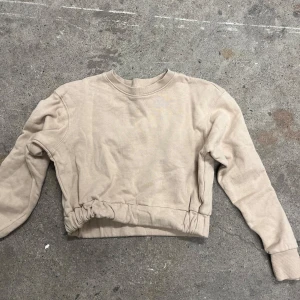 Beige croppad sweatshirt från 157 New - Beige sweatshirt från 157 New med croppad passform och snygg dragsko-detalj i midjan. Tröjan har rund halsringning, långa ärmar och är gjord i mjuk bomullsmix. Perfekt för en chill och trendig look.
