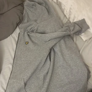 Grå hoodie från Lyle & Scott - Säljer en grå hoodie från Lyle & Scott med dragkedja och huva. Tröjan har det klassiska gula logomärket på bröstet och är tillverkad i mjuk bomull. Perfekt för chill dagar eller när du vill ha en clean och enkel stil.
