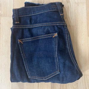 Mörkblå jeans från Nudie Jeans - Snygga mörkblå jeans från Nudie Jeans med klassisk femficksdesign och kontrastsömmar. Jeansen har en rak passform och är tillverkade i slitstarkt denimtyg av bomull. Perfekta för dig som gillar stilrena och tidlösa jeans med en modern touch.