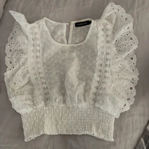 Vit blus med volang  - Supersöt vit blus med volangiga ärmar och smockad midja. Blusen har spetsdetaljer framtill och är ärmlös. Endast andvänd fåtal gånger ,Perfekt för sommaren .