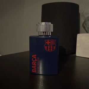 Barça Eau de Toilette 100ml - Parfymflaska i mörkblått med FC Barcelonas logga och text i rött. Flaskan har en robust, fyrkantig form och ett stort, silverfärgat metallock med mönstrad yta. Rymmer 100 ml. Perfekt för dig som älskar fotboll och vill ha en sportig doft.