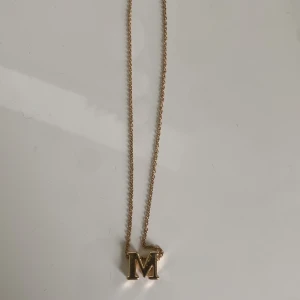 Guldigt halsband med bokstaven M - Snyggt halsband i guldton med en kedja och ett hängsmycke i form av bokstaven M. Kedjan har justerbar längd och ett klassiskt lås. Perfekt accessoar för dig som vill ha något personligt och stilrent.