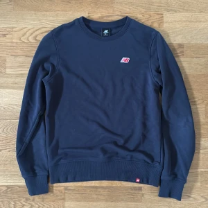 Mörkblå sweatshirt från New Balance - Mörkblå sweatshirt från New Balance med liten logga på bröstet. Klassisk rund halsringning och ribbade muddar vid ärmslut och nederkant. Perfekt för en avslappnad och sportig stil.