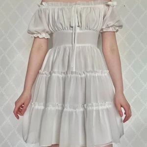 Vit offshoulder kortklänning med volang - Vit kortklänning med offshoulder och puffärmar. Klänningen har volanger i flera lager, smockad rygg och knyt framtill. Så lätt och skön! Perfekt till i sommar eller till studenten🥰Bara använd en gång!