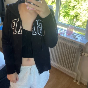 Svart hoodie från GAP - Svart hoodie från GAP med dragkedja och stor vit logga framtill. Koftan har huva, långa ärmar och kängurufickor. Perfekt för chill dagar och enkel att matcha med allt. Mjuk och skön, tillverkad i bomullsmix. Den är mer som xs