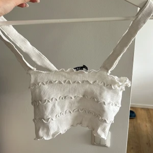 Vit ribbad croppad topp från Zara - Superfin vit croppad topp från Zara med breda axelband och volangdetaljer. Toppen är ribbad och har vågiga sömmar som ger en söt och trendig look. Perfekt till jeans eller kjol när du vill ha en chill men snygg stil.