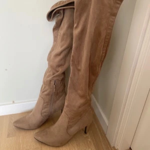 Beige mockastövlar med klack - Snygga knähöga stövlar i beige mocka med spetsig tå och smal klack. Stövlarna har dragkedja på insidan för enkel påtagning och en elegant, slimmad siluett. Perfekta för att lyfta din outfit med en trendig touch.