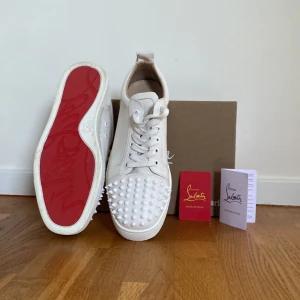 Vita sneakers med nitar från Christian Louboutin - Säljer ett par vita sneakers från Christian Louboutin med ikoniska röda sulor och vita nitar framtill. Skorna har snörning och är tillverkade i skinn. Kommer med originalkartong och äkthetskort. En nit saknas, inget som syns men därav priset
