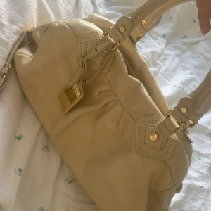 Beige handväska från Marc by Marc Jacobs - Snygg vit handväska i skinn från Marc by Marc Jacobs med guldfärgade metalldetaljer och dubbla handtag. Väskan har en klassisk form, dekorativ bricka framtill och ett innerfoder med svartvitt logomönster. Perfekt för dig som gillar stilrena accessoarer. Jättebra stick. Storlek medium. 