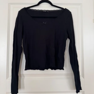 Svart långärmad topp med spets från BikBok - Svart långärmad topp från BikBok med ribbad struktur, söt liten rosett framtill och spetskant runt den breda urringningen. Modellen är figurnära och har vågig kant nertill för en extra detalj. Perfekt för dig som gillar en enkel men ändå söt stil.