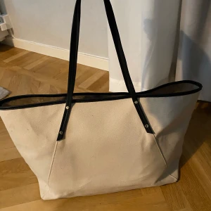 Beige shoppingväska från H&M - Stor och rymlig shoppingväska från H&M i beige bomullstyg med svarta detaljer och långa handtag i syntet. Perfekt för att bära med sig allt man behöver till skolan, stranden eller stan. Enkel och stilren design med innerficka för småsaker. 54 cm bred och 30 cm hög 