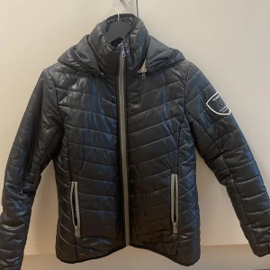 Svart dun rid/stall jacka  Jacson  - Svart rid/stall pufferjacka från Jacson med quiltad design och huva. Jackan har två dragkedjeförsedda fickor framtill och en snygg badge på ärmen. Perfekt för kalla dagar och riktigt skön att bära. Stängs med dragkedja hela vägen upp. 