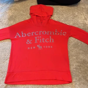 Röd hoodie från Abercrombie Kids - Snygg röd hoodie från Abercrombie Kids med stort silverfärgat tryck på bröstet. Tröjan har huva, långa ärmar och är gjord i mjuk bomull. Perfekt för dig som gillar en enkel men stilren look med tydlig logga framtill.