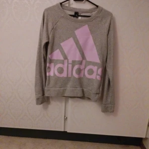 Grå Adidas sweatshirt med rosa tryck XS - Säljer en grå sweatshirt från Adidas i storlek XS. Tröjan har ett stort rosa Adidas-tryck framtill, rund hals och långa ärmar. Perfekt för en sportig och chill stil. Materialet är mjukt och skönt, troligtvis bomullsmix.