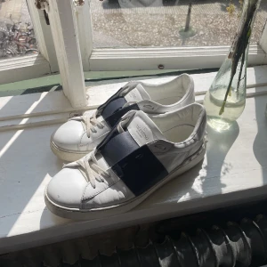 Valentino Open sneakers i vitt och navy - Snygga Valentino Open sneakers i vitt skinn med mörkblått band över mitten och ikoniska nitar på hälen. Perfekta för dig som vill ha en stilren och lyxig look. Bra skick, har endast använt några få gånger. Byter endast mot Golden Goose sneakers i storlek 43!